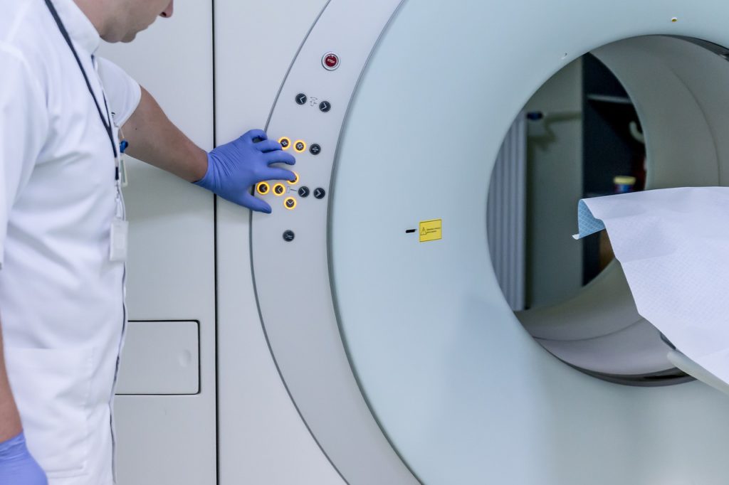 Medical professional wearing gloves operating an MRI scanner control panel beside the scanning chamber in a clinical radiology room.